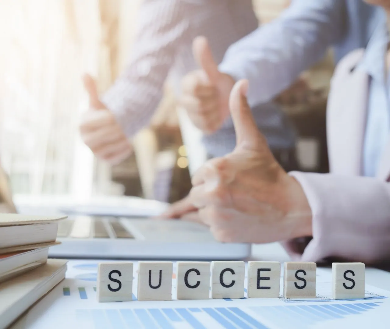 Close-up of scrabble tiles spelling "SUCCESS" on a desk, with two people in the background giving thumbs-up gestures, representing a commitment to achieving business goals through effective SEO strategies for real estate agents.