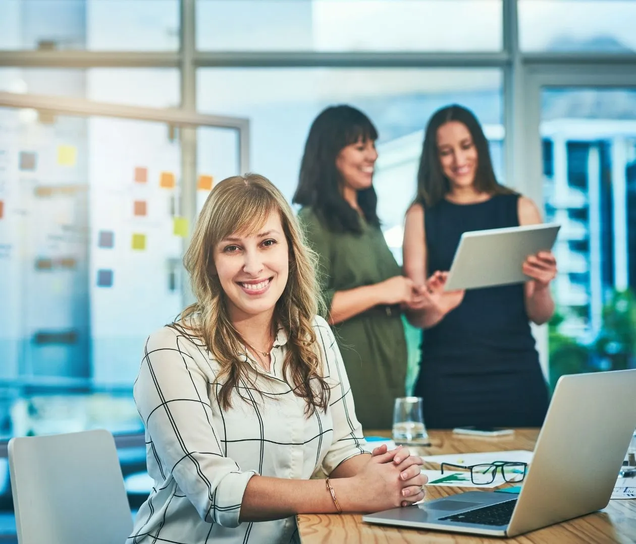 Smiling woman at a desk with a laptop, representing an experienced SEO expert, with two colleagues in the background discussing strategies, highlighting expertise in website optimization for lawyers and law firms.