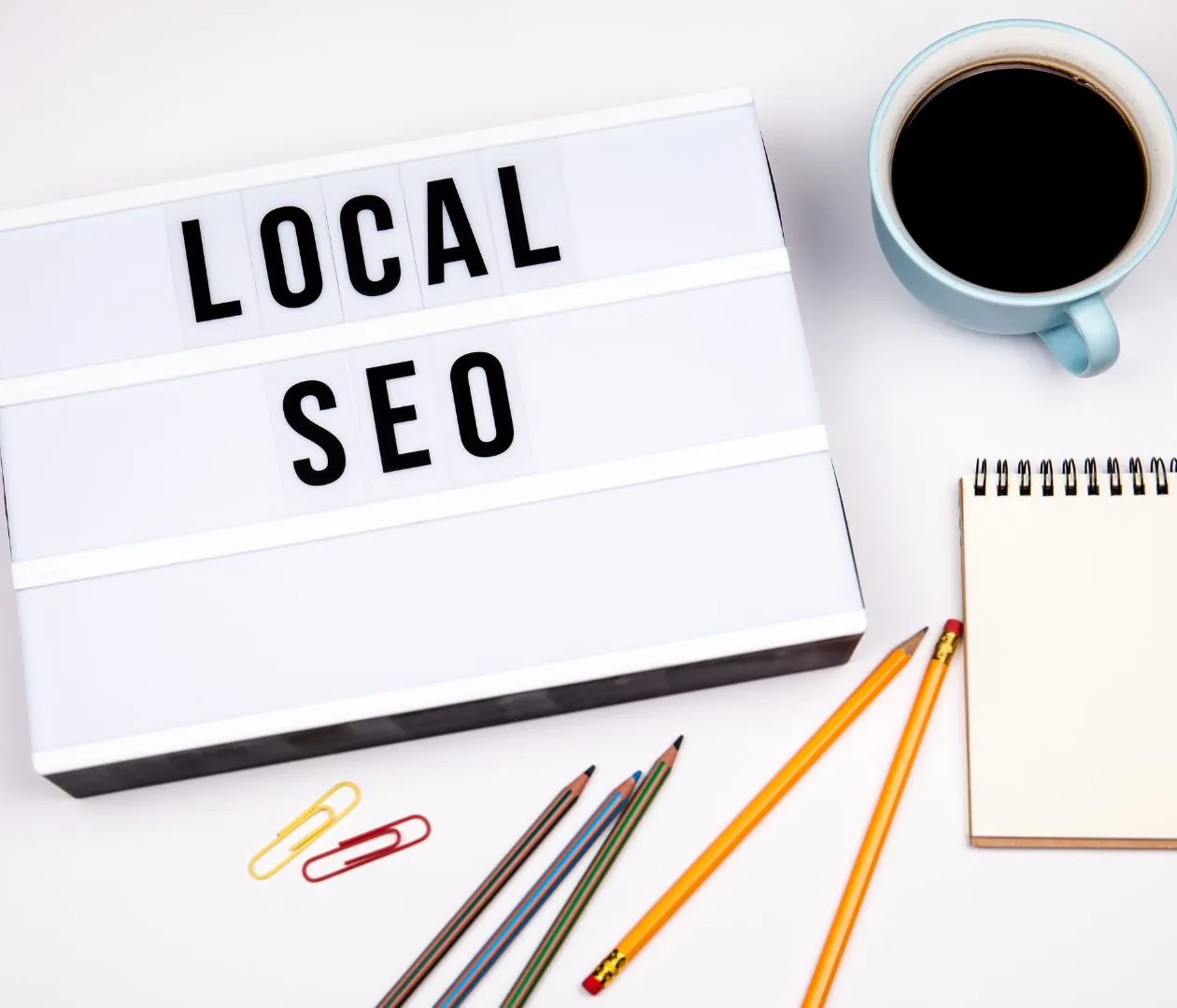 Lightbox sign displaying the words "Local SEO" next to a notebook, pencils, paper clips, and a cup of coffee, symbolizing the importance of local keyword optimization for real estate agents to improve search engine rankings.